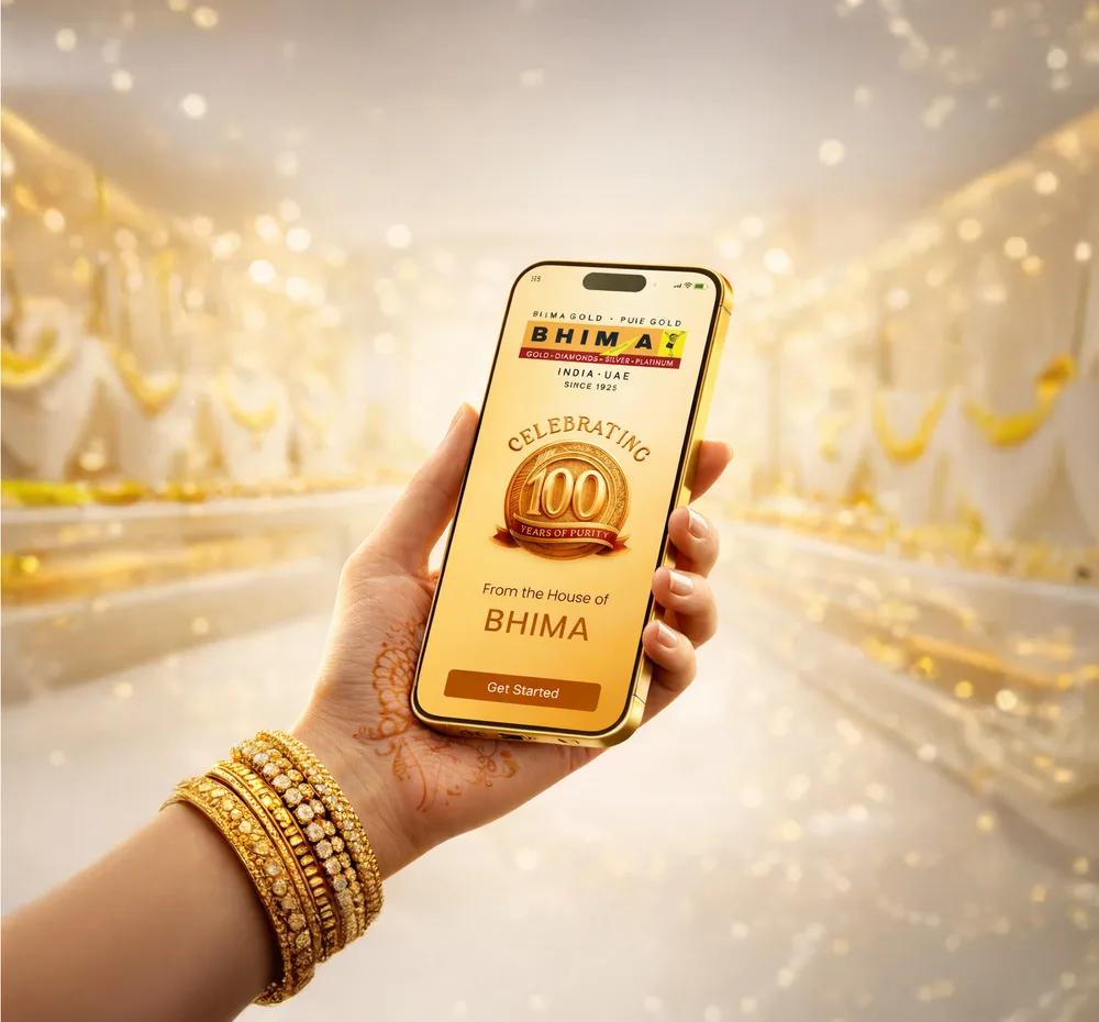 A hand holding a phone showing the Bhima E-Gold app amid golden bokeh